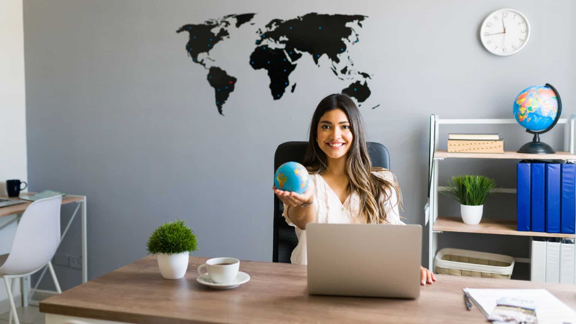 Mujer profesional sonriente en su oficina con un globo terráqueo, representando las oportunidades de convertirse en agente de seguros con alcance global.